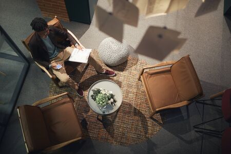 High angle view of young African businessman sitting on chair alone and reading a business contract at fashionable officeの写真素材