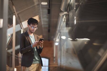 African young businessman in eyeglasses typing a message on his mobile phone while standing at office corridorの写真素材