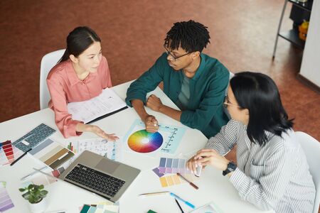 Multiethnic group of graphic designers sitting at the table with colored palette and choosing the color during their work at officeの写真素材