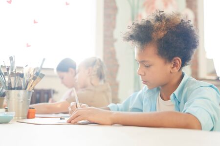 African little boy drawing at the table with other children in the background during art lessonの写真素材