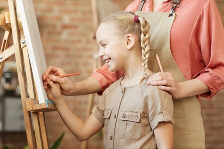 Smiling little girl standing in front of easel and learning to paint with the help of teacher at art studioの写真素材