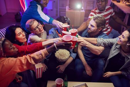 Group of young people toasting with beer while sitting at home with popcorn and they are going to...