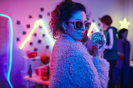 Portrait of young party girl in sunglasses and in stylish jacket looking at camera while dancing at...