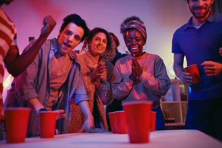African smiling woman is very excited while playing in beer pong game together with her friends...