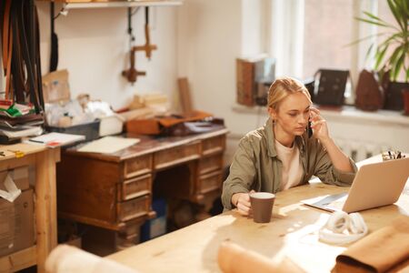 Horizontal high angle shot of young woman wearing casual outfit working on laptop talking on phone...