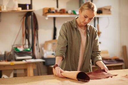 Young woman wearing casual clothes standing at table in her workshop rolling out piece of brown...