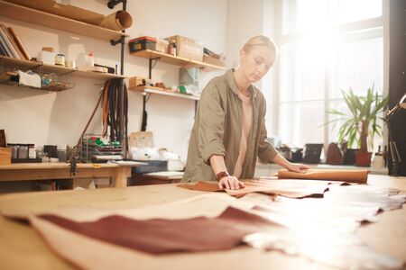 Young woman standing at table in her workshop rolling out leather material for new craftwork,...