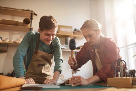 Young adult female artisan using hammer and punching leather tool while her young male colleague...
