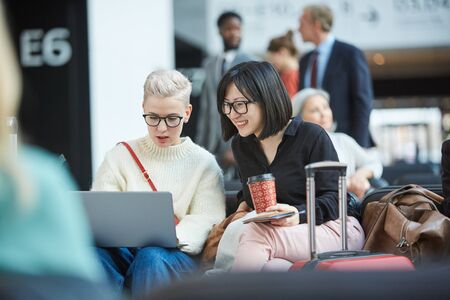 Two young female friends sitting together in departure lounge watching something funny on laptop,...