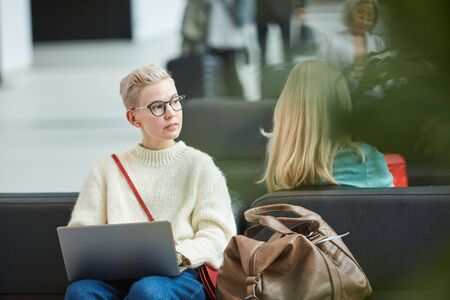 Horizontal medium portrait of young blond woman wearing eyeglasses sitting in departure lounge looking away while surfing Internet on her laptopの写真素材
