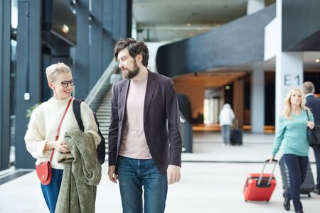 Young Caucasian man and woman chatting while walking along airport hall, horizontal medium long shot