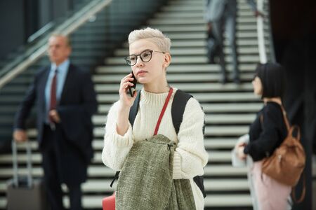 Horizontal portrait shot of stylish young woman standing in airport hall talking on phone, copy...