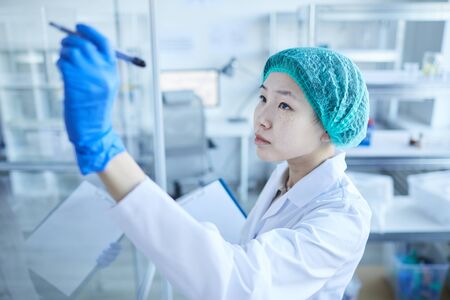 Horizontal medium close-up high angle shot of female Asian laboratory scientist writing formulae on glass wallの写真素材
