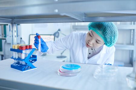 Horizontal medium close-up shot of young female Asian laboratory worker taking out test-tube with...