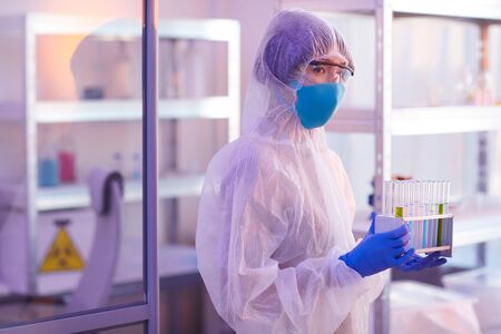 Unrecognizable chemical industry worker wearing disposable protective lab uniform holding test-tubes...