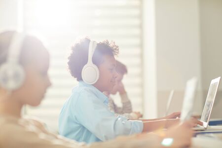 Medium side view portrait of modern twelve-year-old Black boy wearing headphones using laptop during...