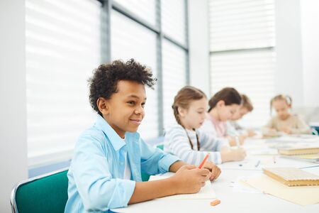 Horizontal portrait of five children wearing casual outfits sitting at desk in modern classroom...