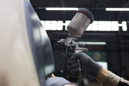 Auto service worker's hand in protective glove holding spay-gun painting car body, horizontal close...