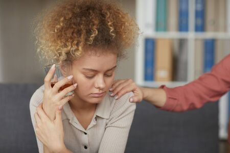 Sad young woman sitting on sofa and drinking whiskey she has difficulties and her friend consoling herの写真素材
