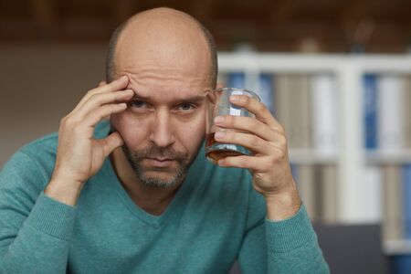 Portrait of drunk mature man holding glass with whiskey and looking at camera with sad sightの写真素材