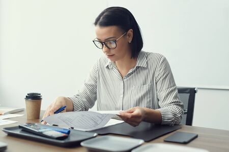 Young businesswoman in eyeglasses sitting at the table with coffee cup and doing paperwork at officeの写真素材