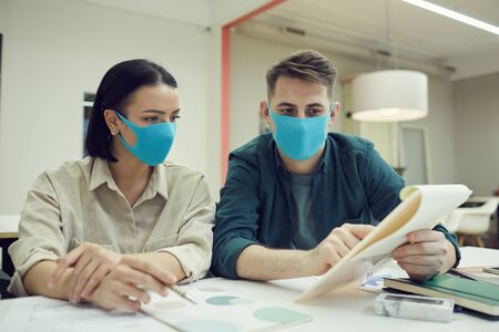Two business colleagues wearing protective masks and discussing business document together at the table at officeの写真素材