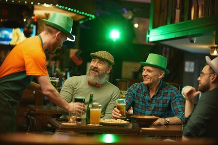 Group of men sitting at the table and talking to the waiter while he bringing glasses of beer for...