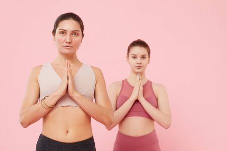 Portrait of two beautiful girls standing in yoga pose and looking at camera isolated on pink backgroundの写真素材