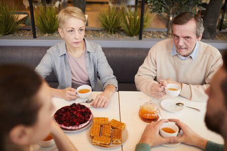 Mature parents sitting at the table drinking tea with cakes and talking to their children during breakfast in cafeの写真素材