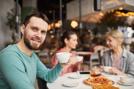 Portrait of young man smiling at camera and drinking tea with his family during lunchの写真素材