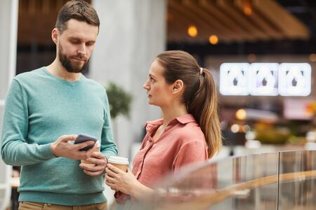Young woman drinking coffee and talking to man while he using his mobile phone they are in the mall