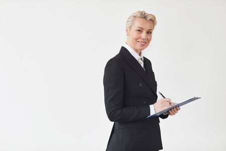 Portrait of mature businesswoman in black suit making notes in document and smiling at camera over...