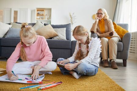 Two sisters sitting on the floor and drawing with colored pencils with their grandmother sitting in the backgroundの写真素材