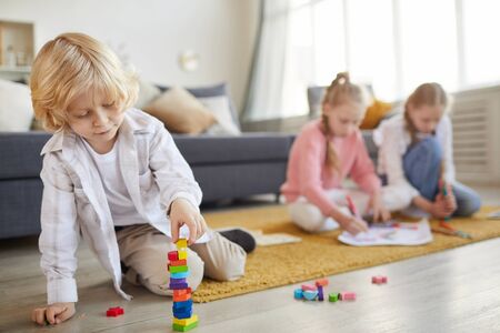 Little boy with blond hair playing on the floor with toys with his two sisters drawing in the background in the living roomの写真素材