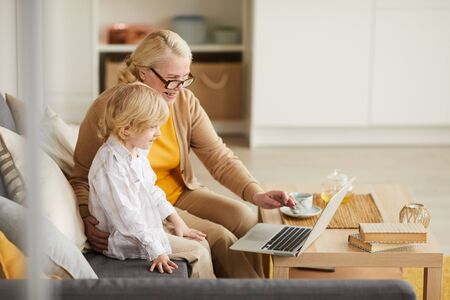 Happy mature woman sitting on sofa with her grandson and they using laptop for online conversation with his parentsの写真素材