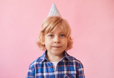 Portrait of little boy with blond hair and in party hat looking at camera over pink background