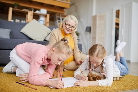 Two little girls lying on the floor and drawing with their grandmother in the background they are at homeの写真素材