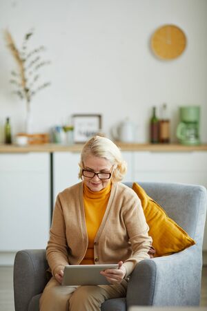 Mature woman in eyeglasses sitting n armchair and using digital tablet in the living room at homeの写真素材
