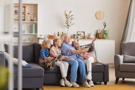 Grandmother sitting on sofa together with her grandchildren and they making a selfie on mobile phone...
