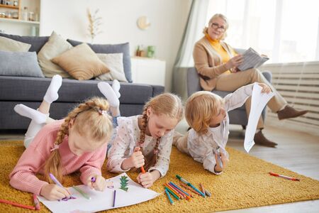Little girls and boy lying on the floor and drawing with colored pencils with their grandmother reading a book in armchair in the backgroundの写真素材