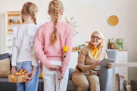 Smiling grandmother sitting on sofa and looking at her granddaughters how they congratulating her with holidayの写真素材