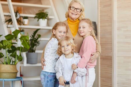 Portrait of happy grandmother in eyeglasses embracing her grandchildren and smiling at camera while...