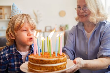 Happy little boy blowing candles on his birthday cake together with his grandmother at homeの写真素材