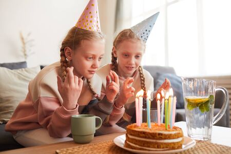 Two cute girls in party hats eating birthday cake at the table in the living room at home