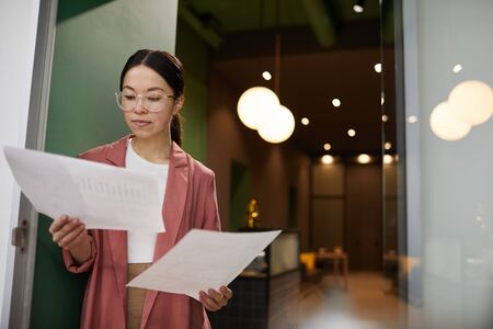 Young Asian businesswoman in eyeglasses examining future project on papers while standing at office