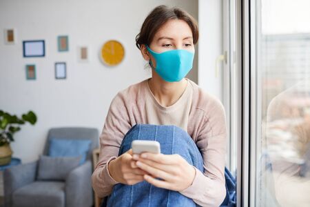 Young woman wearing protective mask she is ill and sitting at home she using her mobile phone and...