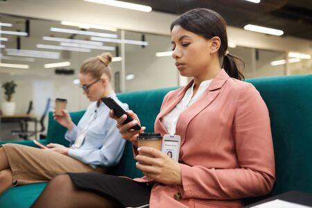 Serious African businesswoman sitting on sofa and waiting for the meeting she drinking coffee and using her mobile phoneの写真素材