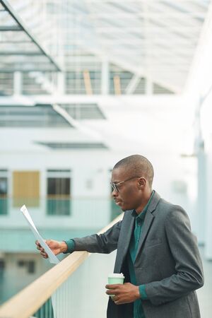 African young businessman drinking coffee and reading a document while standing at office corridorの写真素材