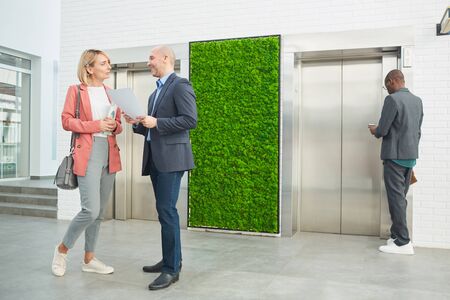 Business people talking to each other while standing at office corridor and waiting for the elevator