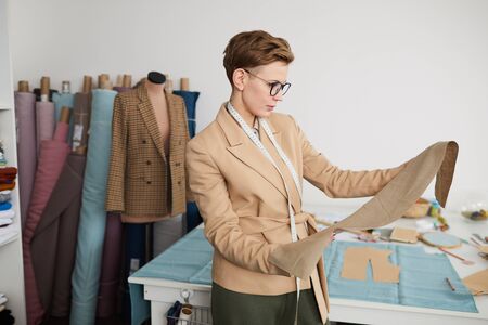 Serious tailor in eyeglasses holding fabric in her hands and examining it before work